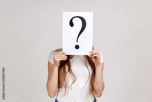 Portrait of anonymous woman hiding her face behind white paper with question mark, finding smart solution, asking for advice, wearing white T-shirt. Indoor studio shot isolated on gray background.