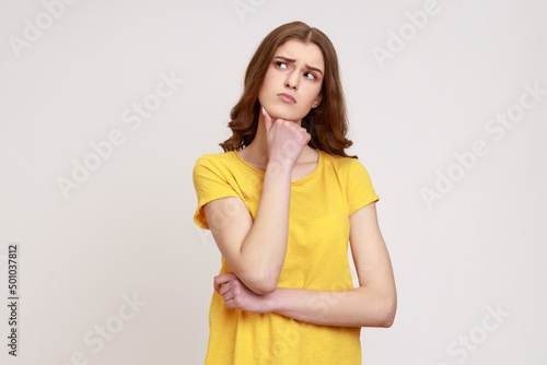 Portrait of attractive teenager girl with brown wavy hair in yellow T-shirt thinking about important things  holding chin  looking away. Indoor studio shot isolated on gray background.