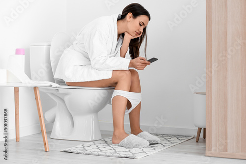 Young woman using mobile phone on toilet bowl in restroom