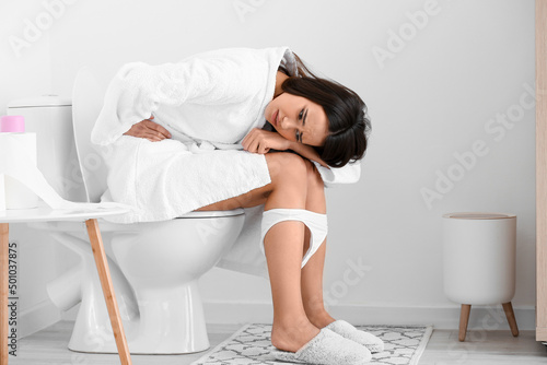 Fotografie Sad young woman suffering from bellyache on toilet bowl in restroom