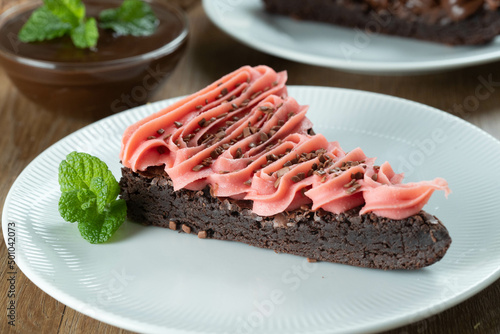 Piece of Chocolate Brownie Slice with Strawberry and Chocolate Icing. Wooden table with mint and chocolate chips in the background