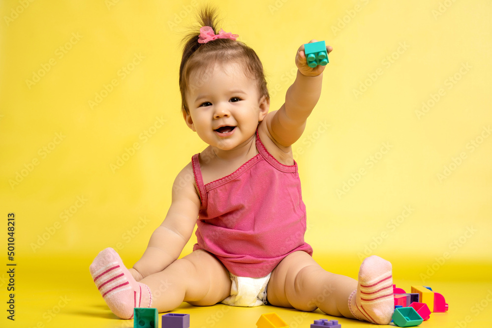 A 1 years old girl is playing with a children's multi-colored constructor. The girl is sitting on a yellow background in a diaper. A 1 years old girl is playing with a children's multi-colored constructor. The girl is sitting on a yellow background in a diaper.