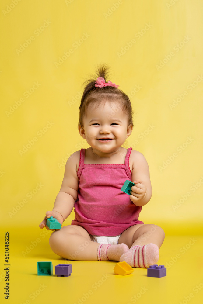 Naklejka premium A 1 years old girl is playing with a children's multi-colored constructor. The girl is sitting on a yellow background in a diaper.