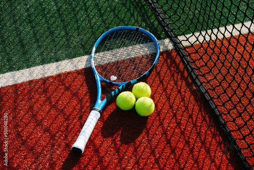 Tennis racket and three standart balls with net shadow cast on it. On a brand new outdoor court. Vibrant green and brown with clear white lines. © zzzdim