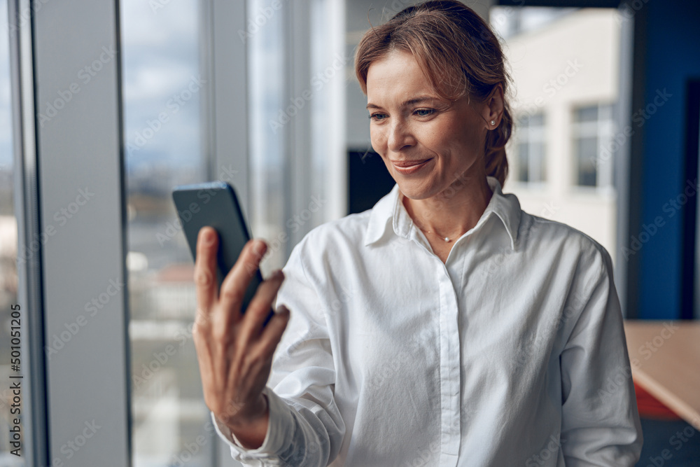 Smiling woman standing in office. Businesswoman with mobile phone in hand looking at smartphone.