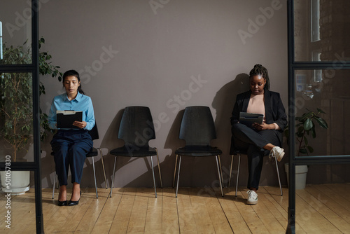 Two women sitting on chairs in hallway and filling form before job interview during pandemic