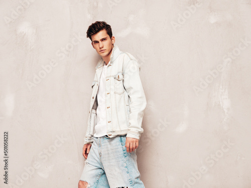 Portrait of handsome confident stylish hipster lambersexual model.Man dressed in jacket and jeans. Fashion male posing in studio near grey wall