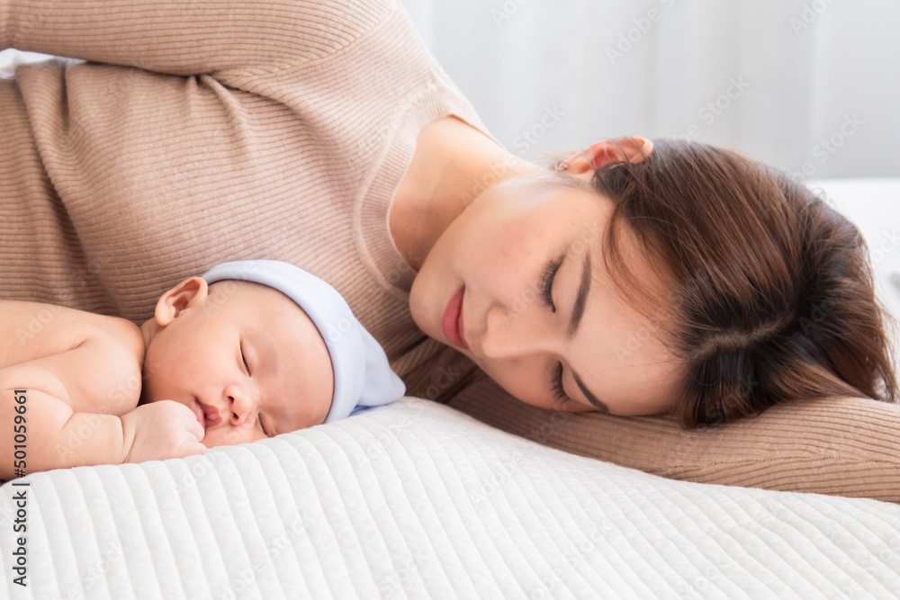 Selective focus Asian mother lying on bed with newborn baby wearing blue hat in home, mom hugging little baby with love and care. Woman carying her adorable infant while toddler sleeping in bedroom. Selective focus Asian mother lying on bed with newborn baby wearing blue hat in home, mom hugging little baby with love and care. Woman carying her adorable infant while toddler sleeping in bedroom.