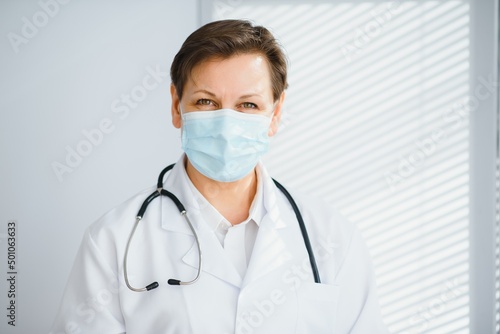 Older female doctor wearing face mask and white medical coat standing in hospital. Portrait