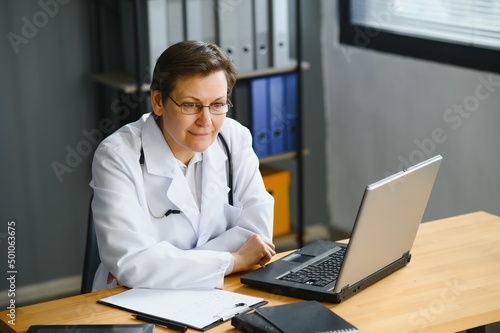 Portrait shot of middle aged female doctor sitting at desk and working in office.