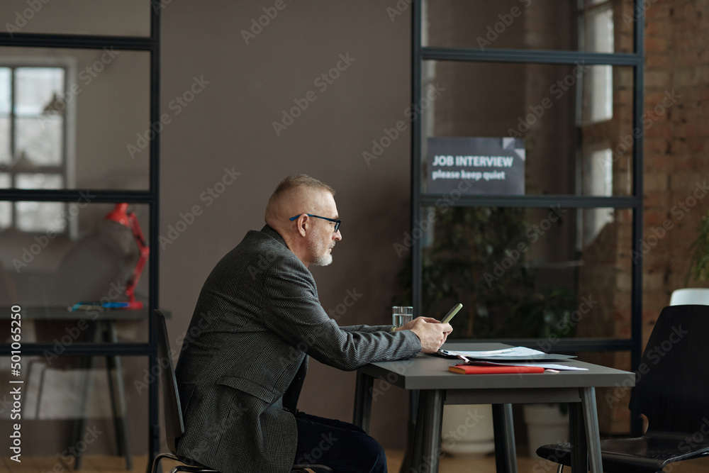 Mature employee in eyeglasses sitting at table and using mobile phone while waiting for job interview at office Mature employee in eyeglasses sitting at table and using mobile phone while waiting for job interview at office