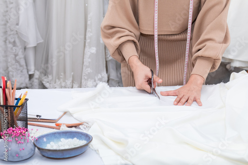 Selective focus close up of hand working woman cutting fabric with scissors while sewing custom made clothes and design wedding dress in work shop. Fashion designer work at creative office.