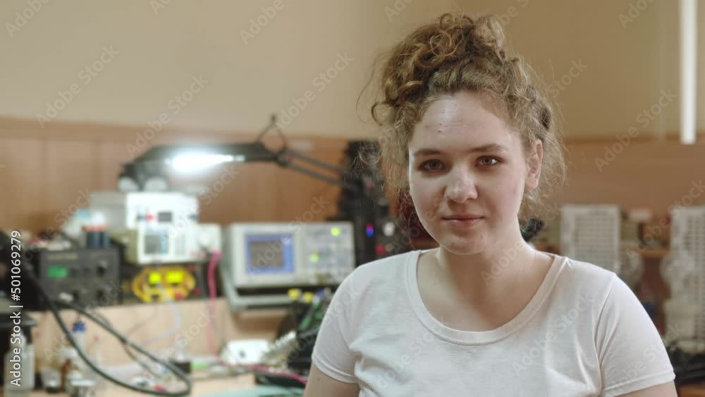 Happy female worker turns and smiles to a camera at electronics repair service workshop. Woman fixes household appliances, computers and electrical equipment in her workroom.