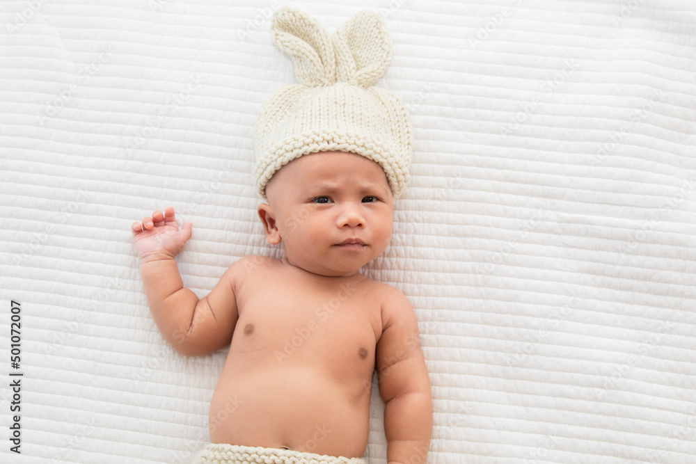 Asian adorable newborn baby wearing pants and rabbit hat beige knitted lying on white bed. little baby 0-1month looking surprised at camera. concept of happy childhood and motherhood. Asian adorable newborn baby wearing pants and rabbit hat beige knitted lying on white bed. little baby 0-1month looking surprised at camera. concept of happy childhood and motherhood.