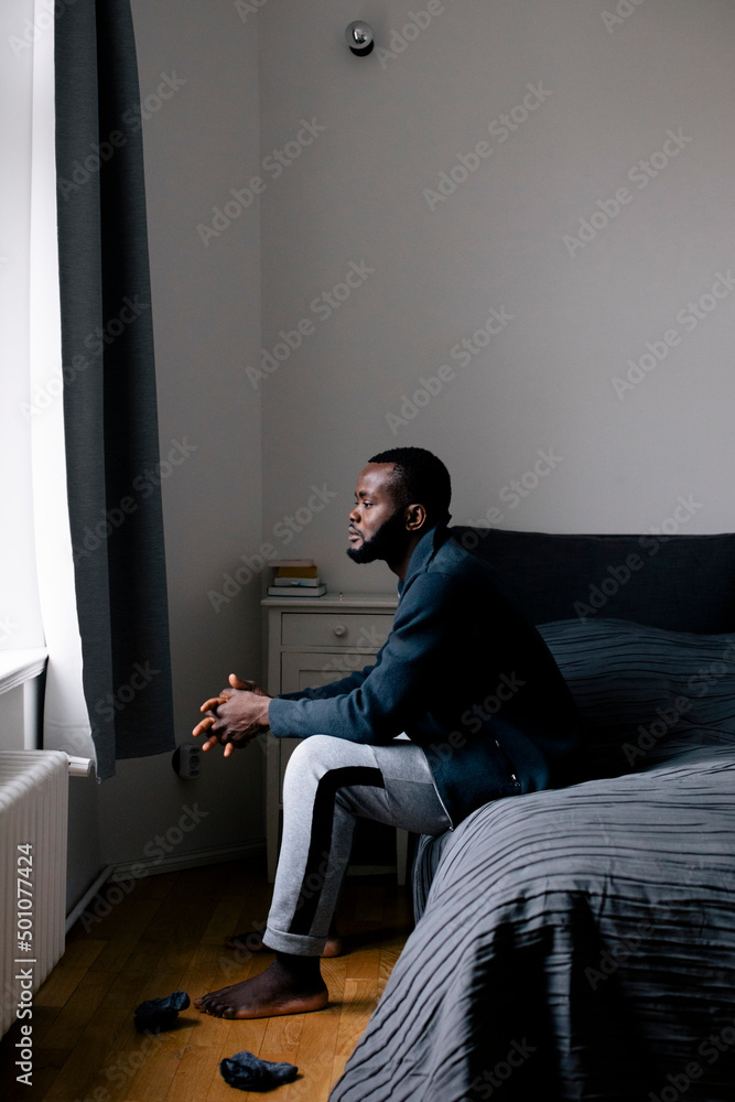 Full length side view of thoughtful man sitting on bed at home Full length side view of thoughtful man sitting on bed at home