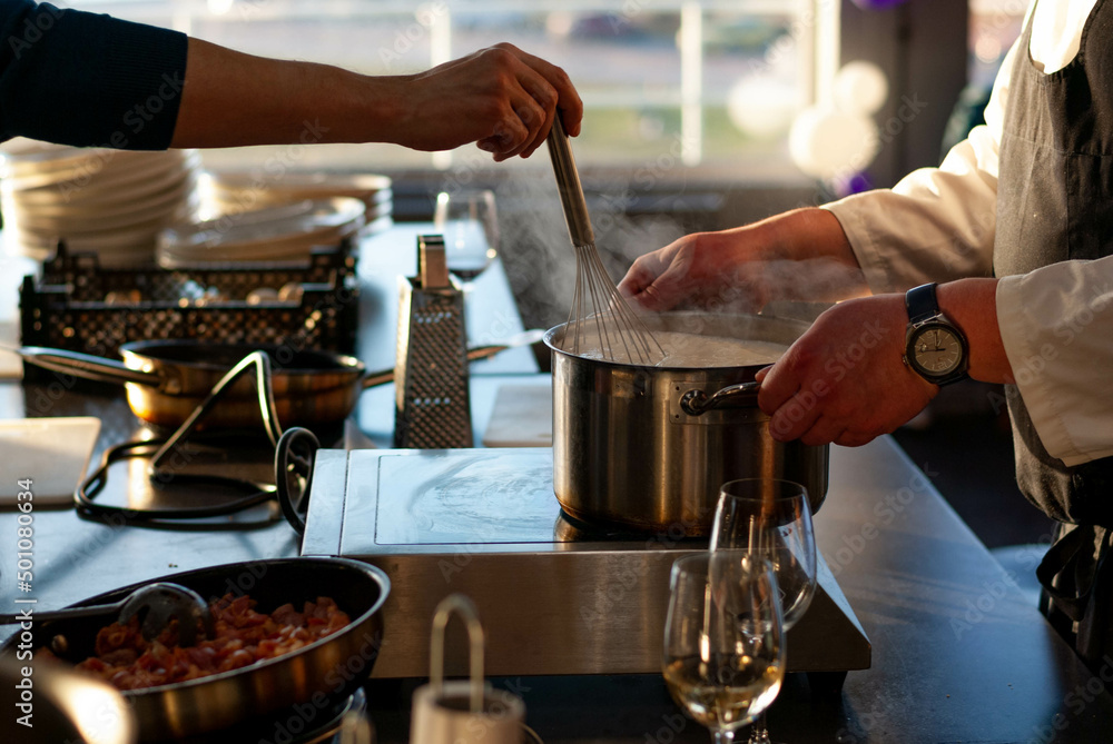in the restaurant kitchen, the chef's hands hold a saucepan over the stove, a woman's hand stirs the dish with a whisk, together they cook a delicious dish