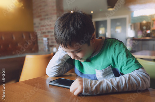 Cinematic portrait young boy playing game on mobile while waiting for food, Kid sitting in coffee shop sending text to friends, Child playing game online on phone.Children with technology concept