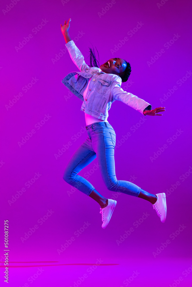 Full body portrait of ecstatic African-American woman jumping in modern neon light studio background
