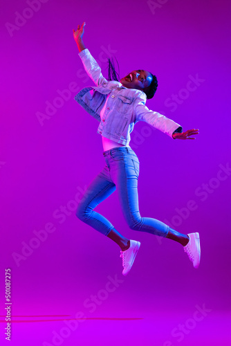 Full body portrait of ecstatic African-American woman jumping in modern neon light studio background