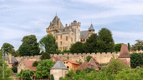 Chateau de Montfort on hill top and village huddled below substantial ramparts of castle with attractive houses in Dordogne region, France