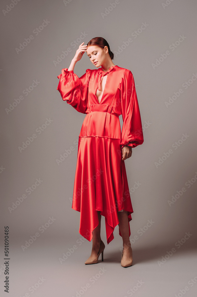 Bright young lady in red oversize dress with long wide sleeves and sexy neckline on high heels mooving on grey studio background. Elegant contemporary bohemian female look. Bright young lady in red oversize dress with long wide sleeves and sexy neckline on high heels mooving on grey studio background. Elegant contemporary bohemian female look.
