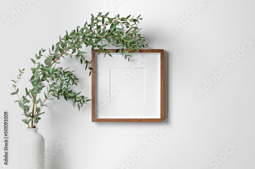 Square frame mockup for artwork, quote or print presentation on white wall with fresh eucalyptus plant in vase photo