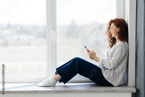 Beautiful red-haired woman sitting on the windowsill in a cozy city apartment. Young girl spends her leisure time using smartphone.