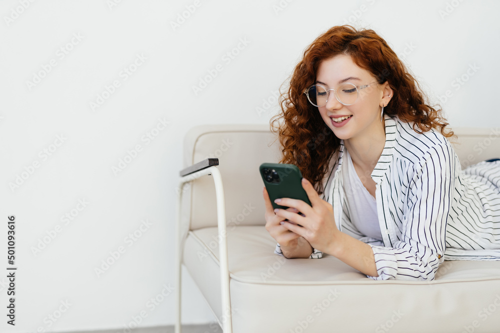 Young red haired woman using smart phone while resting on sofa at home.