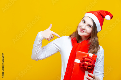 Slika na platnu Winsome Young Caucasian Female Girl Pointing To Giftbox in Santa Hat Over Yellow