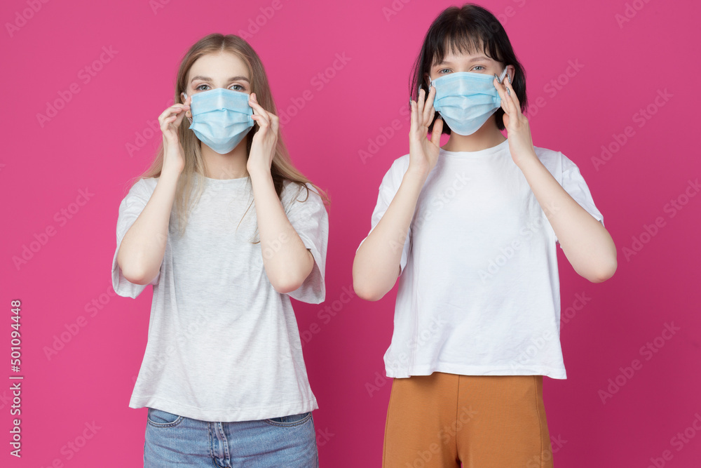 Lifestyle Ideas. Winsome Couple of Two Positive Young Girlfriends Demonstrating Application of Facial Protection Surgical Mask Against Coral Background. Lifestyle Ideas. Winsome Couple of Two Positive Young Girlfriends Demonstrating Application of Facial Protection Surgical Mask Against Coral Background.