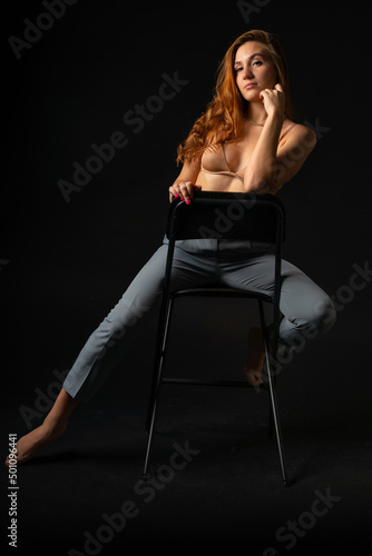Sexy blonde girl model in a pink bra on a bare torso and gray trousers posing barefoot sitting on a chair in the studio on a black background in provocative poses. Seduction in the dark
