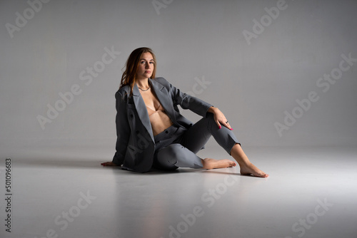 Photos An incredibly beautiful model girl with long blonde hair in an unbuttoned gray jacket, beige bra and gray short trousers is sitting on a floor barefoot