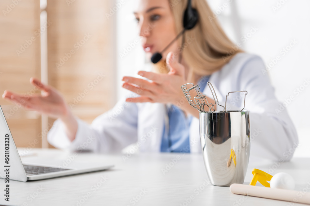 Tools and respiratory muscle trainer near blurred logopedist having video call on laptop in consulting room. Tools and respiratory muscle trainer near blurred logopedist having video call on laptop in consulting room.