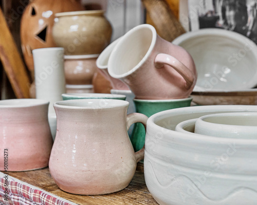 Mugs, plates and other ceramic utensils made of white clay, standing on wood table.
