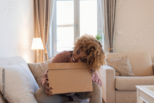 Online shopping, home delivery and satisfied customer. Cheerful interested pretty young african american woman opens cardboard box and looks inside, sits on sofa in living room interior, free space..