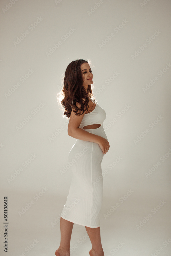 glamour pregnancy. young curly brunette girl in white slim dress stands happy with smile in profile with hands on belly on a white studio wall background in backlight. pregnant concept, free space glamour pregnancy. young curly brunette girl in white slim dress stands happy with smile in profile with hands on belly on a white studio wall background in backlight. pregnant concept, free space