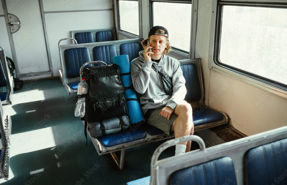 Positive male tourist in casual clothes sits on a bench in a train with a backpack and talks on the phone with a smile on his face.