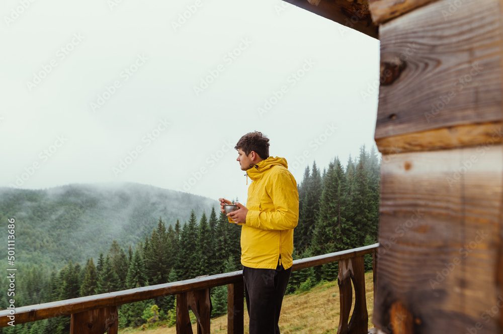 A male tourist stands on the terrace of a house in the mountains with a plate in his hands and looks at the beautiful forest landscape.