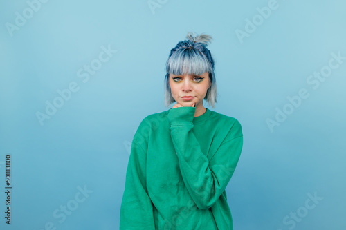Pensive woman in green sweatshirt and with blue hair isolated on blue background with serious face.