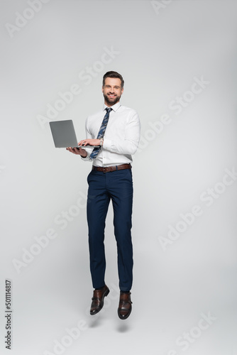 full length of cheerful economist in formal wear levitating with laptop isolated on grey.