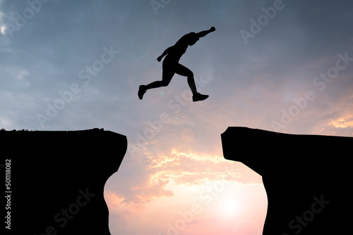 Silhouette young man jumping over precipice between two rocky mountains at sunset.
