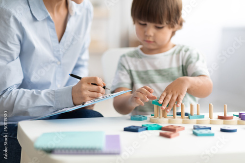 Little boy playing logical wooden toy at psychologist office, woman specialist taking notes, selective focus, free space