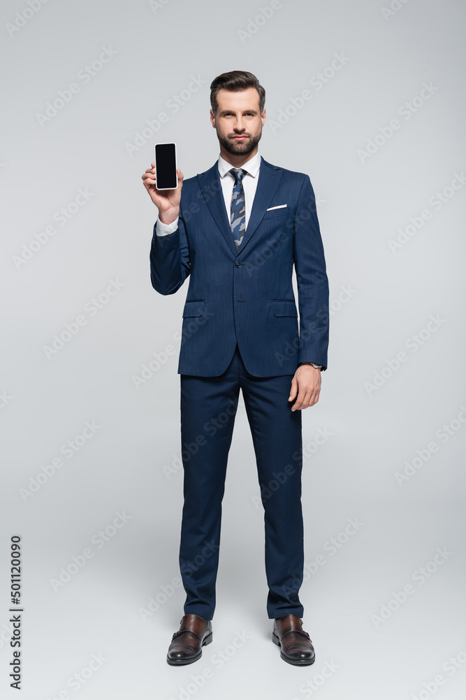full length view of businessman in blue suit showing smartphone with blank screen on grey. full length view of businessman in blue suit showing smartphone with blank screen on grey.