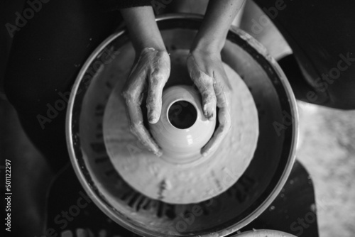 Female hands sculpt clay dishes. Сraftswoman in apron sitting at pottery wheel and using craft tool while shaping wet clay vessel