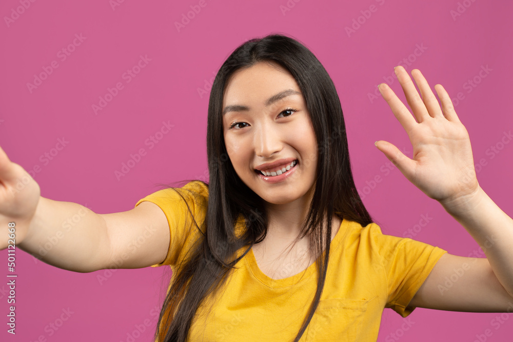 Hello. Happy korean lady taking selfie and waving hand at camera, gesturing hi while making photo over violet background Hello. Happy korean lady taking selfie and waving hand at camera, gesturing hi while making photo over violet background