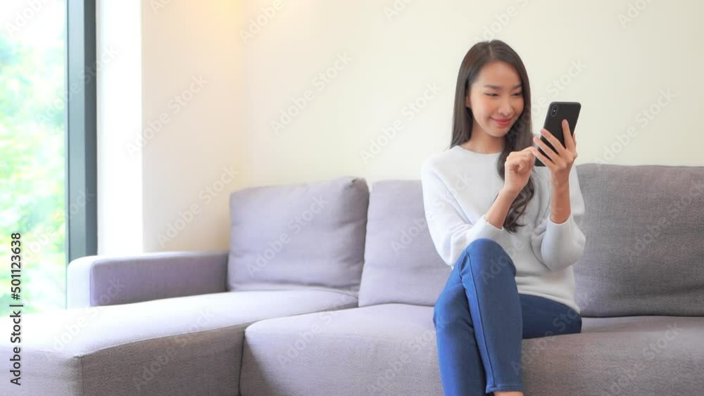 A young attractive woman in jeans and a sweater sits on a couch as she types information into her smartphone. Title space A young attractive woman in jeans and a sweater sits on a couch as she types information into her smartphone. Title space
