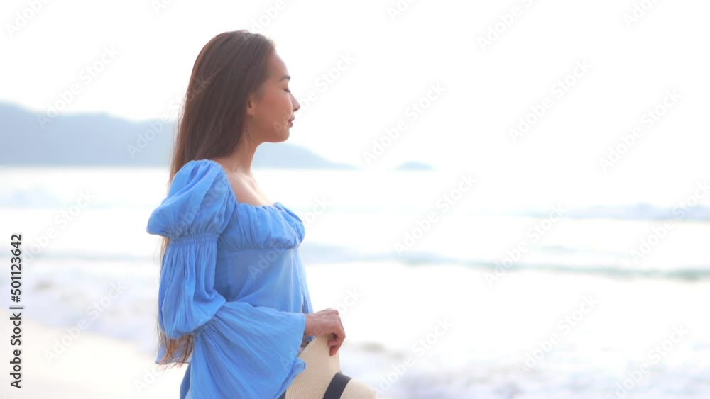 Slow motion view of beautiful Asian woman standing on island beach in blue dress holding hat looking out to sea. Lady enjoying seaside view on tropical vacation holiday. Slow motion view of beautiful Asian woman standing on island beach in blue dress holding hat looking out to sea. Lady enjoying seaside view on tropical vacation holiday.