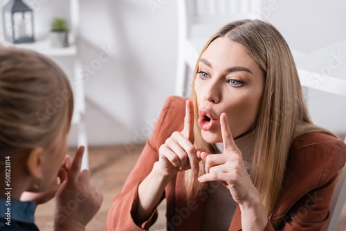 Speech therapist gesturing and talking near blurred girl in classroom.