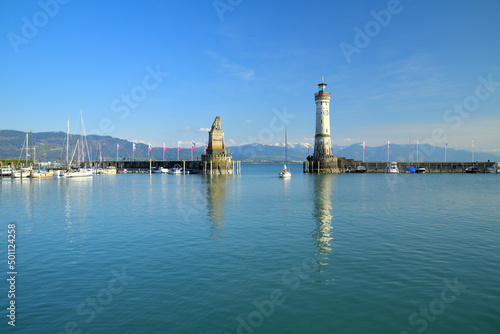 Ein Segelboot fährt gerade bei blauem Himmel aus dem Hafen von Lindau am Bodensee, mit Blick auf die noch schneebedeckten Berge.