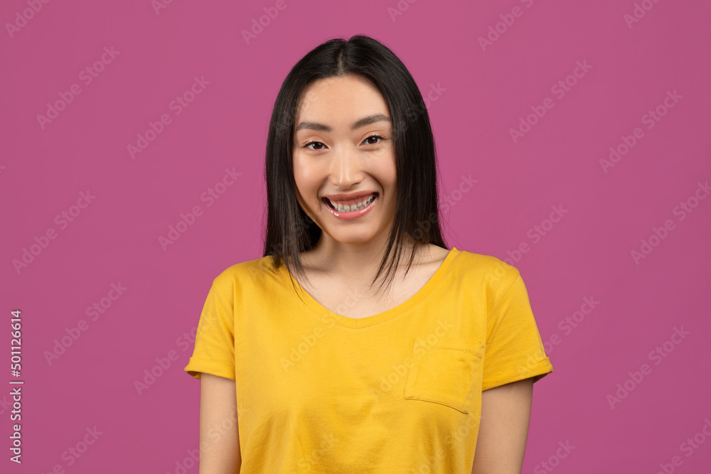 Portrait of cute asian woman in yellow t-shirt posing over violet studio background, looking at camera and smiling Portrait of cute asian woman in yellow t-shirt posing over violet studio background, looking at camera and smiling